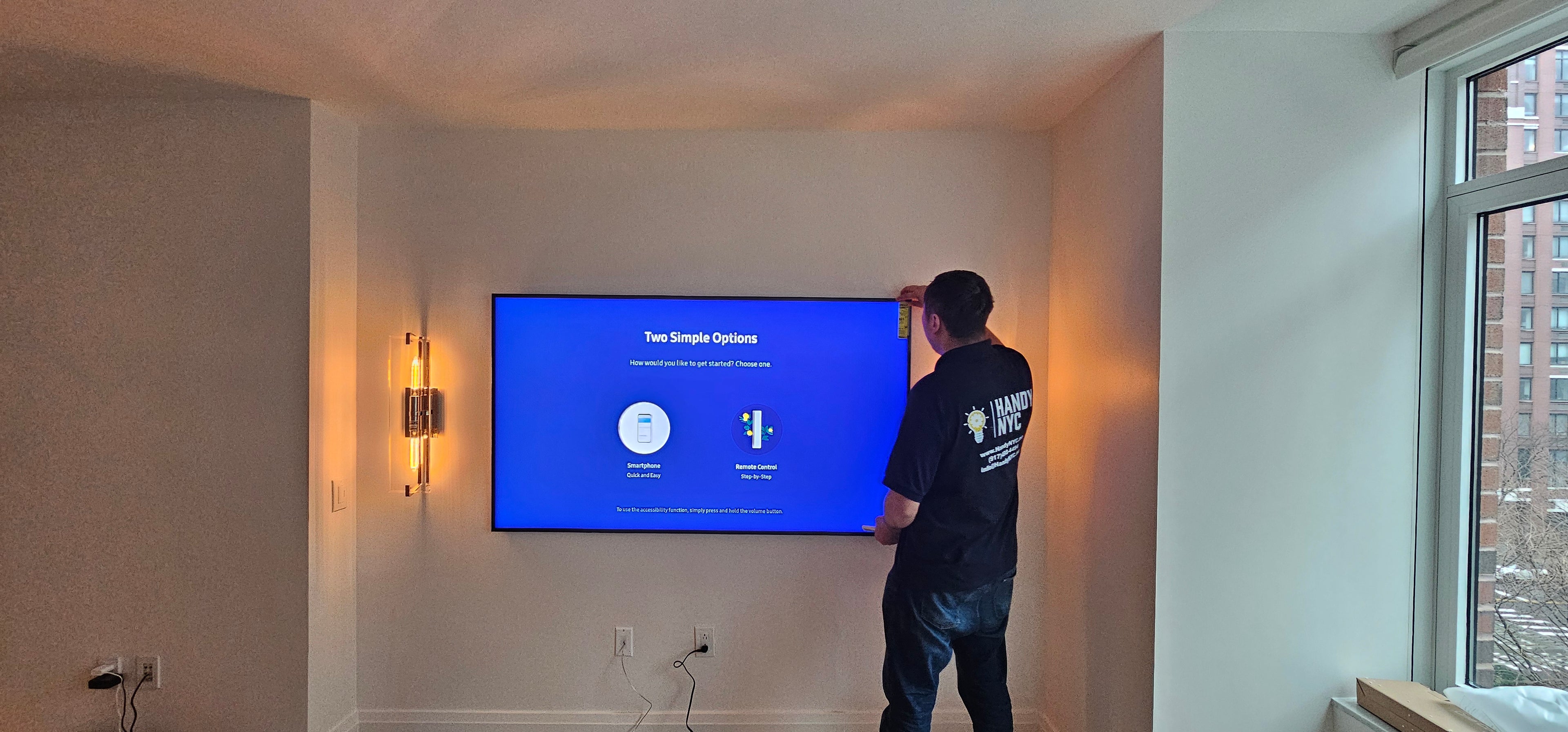 A handyman in HandyNyc shirt taking off protective film off a new tv mounted on the wall.